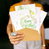 Felicitare scrisă de mână, plic și un stilou pe o masă, alături de câteva flori